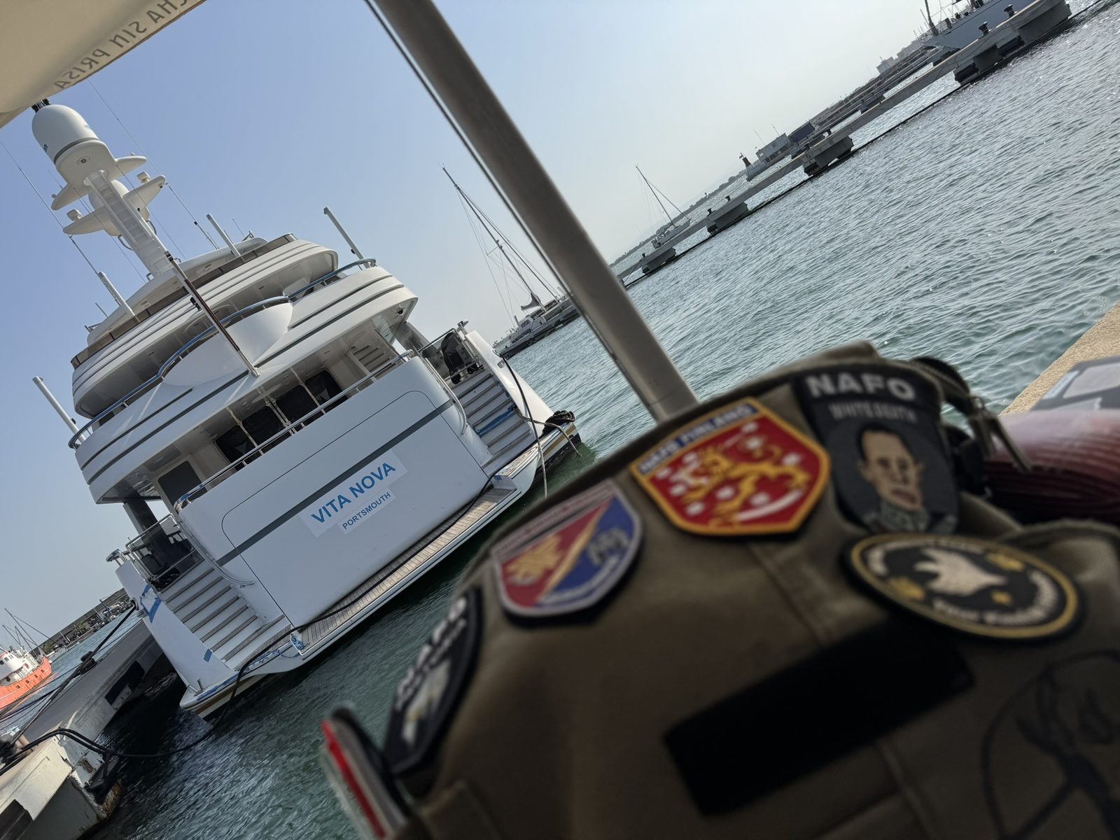 A canvas bag covered in NAFO patches in the foreground. Behind it, moored in the marina, a large white motor yacht named Vita Nova, Portsmouth.
